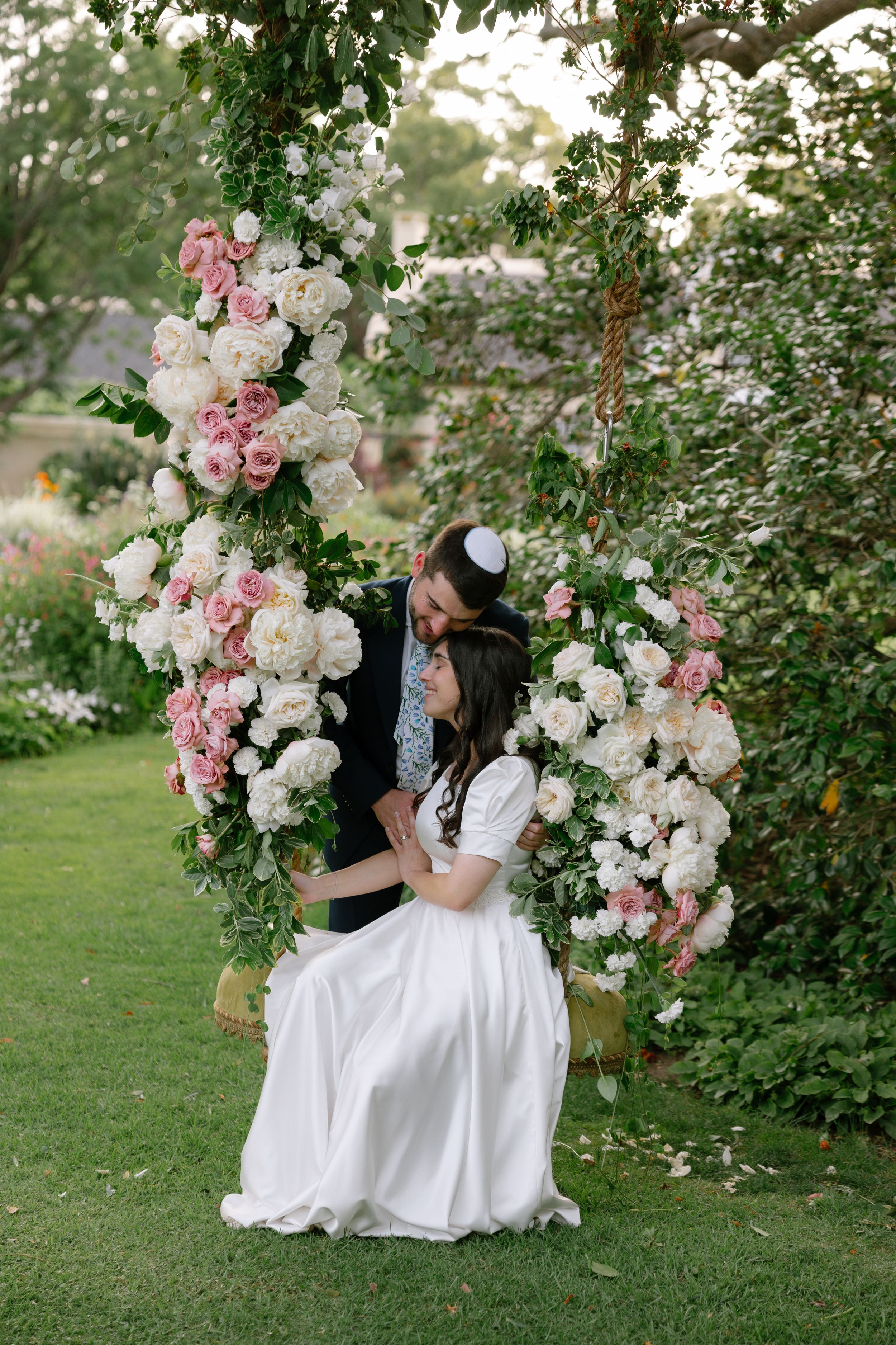 A romantic garden scene with a couple embracing under a floral arch of pink and white roses on a lush green lawn.