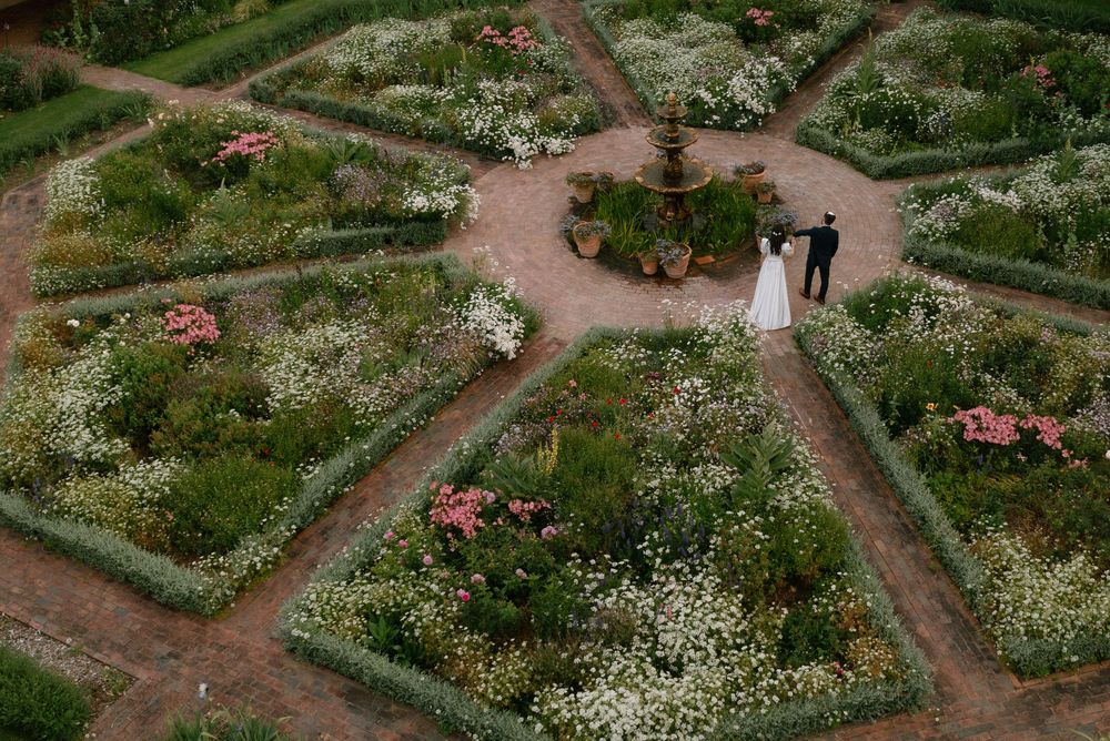 An aerial view shows a garden with intersecting pathways lined with white flowers and green plants.