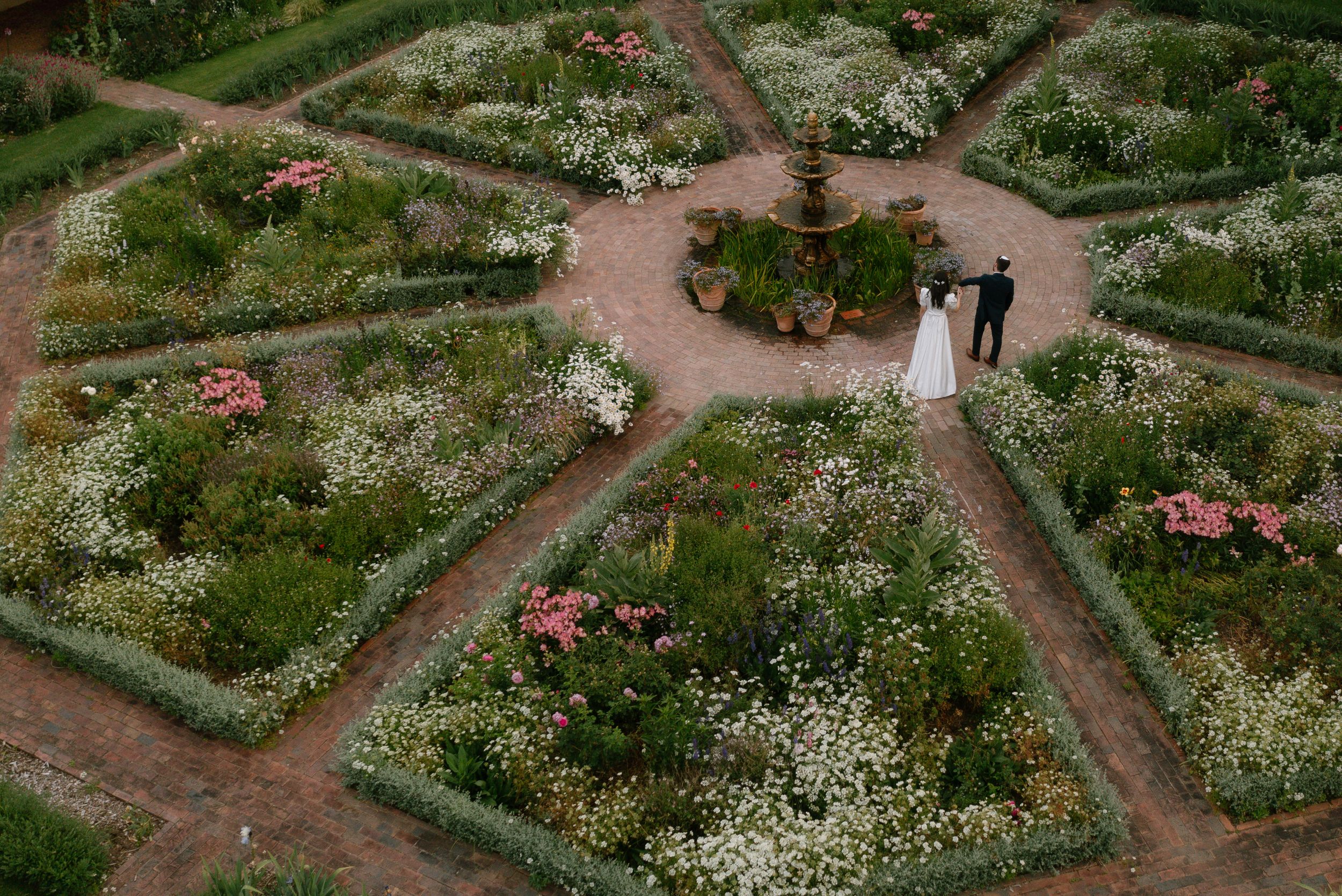 An aerial view shows a garden with intersecting pathways lined with white flowers and green plants.