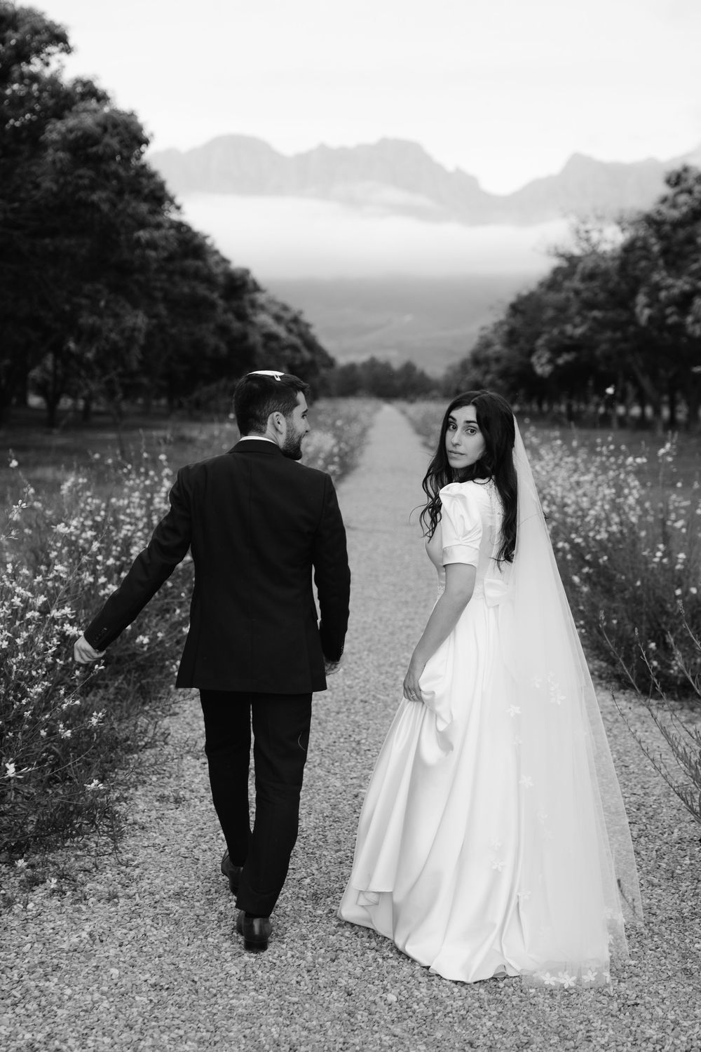 Black and white wedding photo showing couple walking on rural path with mountains and wildflowers in background.