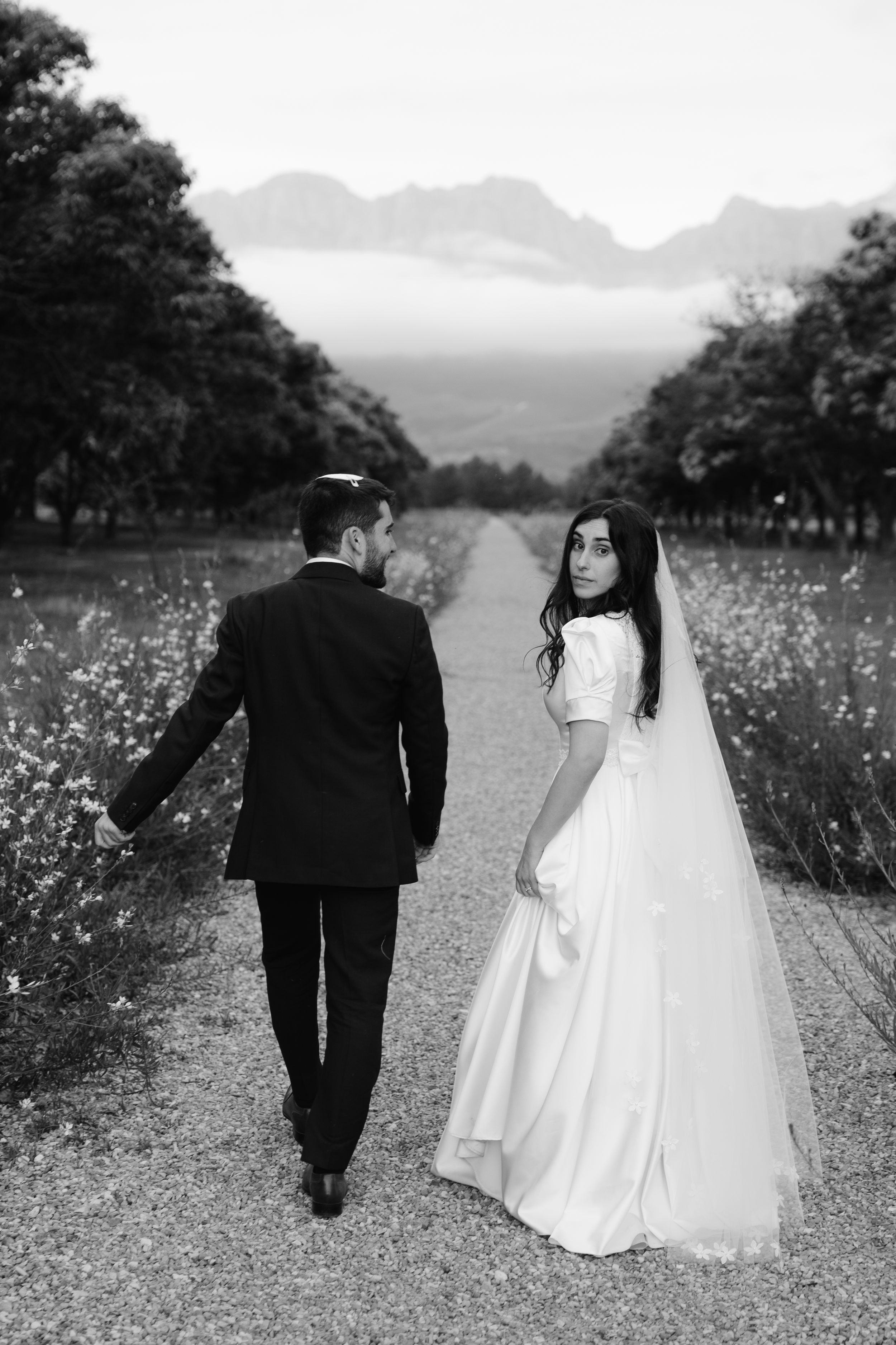 Black and white wedding photo showing couple walking on rural path with mountains and wildflowers in background.