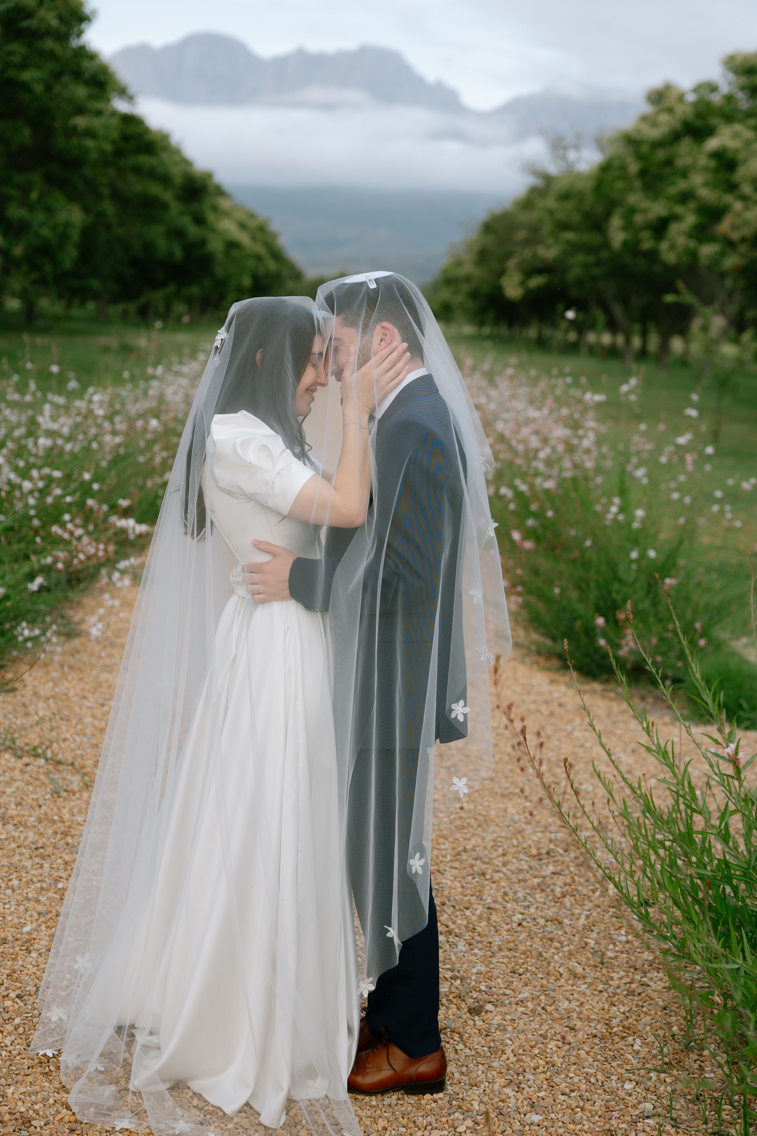 A romantic wedding couple shares an intimate moment on a gravel path with misty mountains in the background.