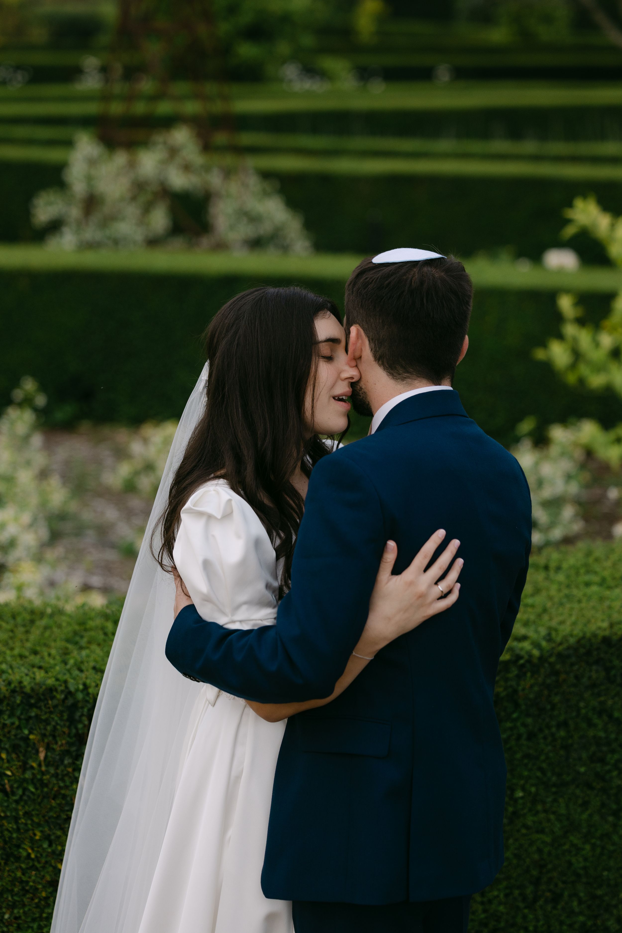 A romantic wedding couple embraces in a garden setting with manicured hedges in the background.