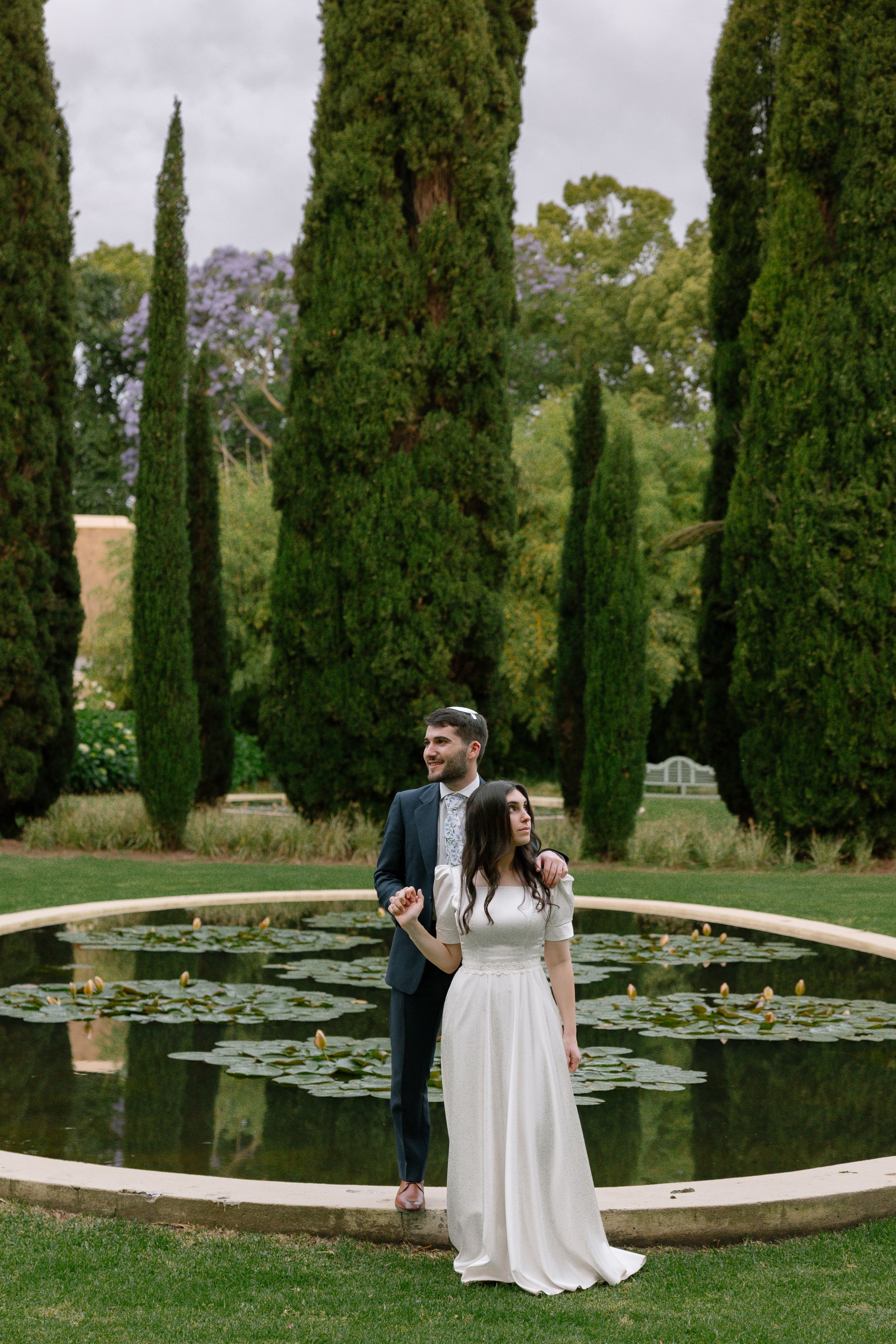 A couple stands by a circular lily pond surrounded by tall cypress trees in an elegant garden setting.