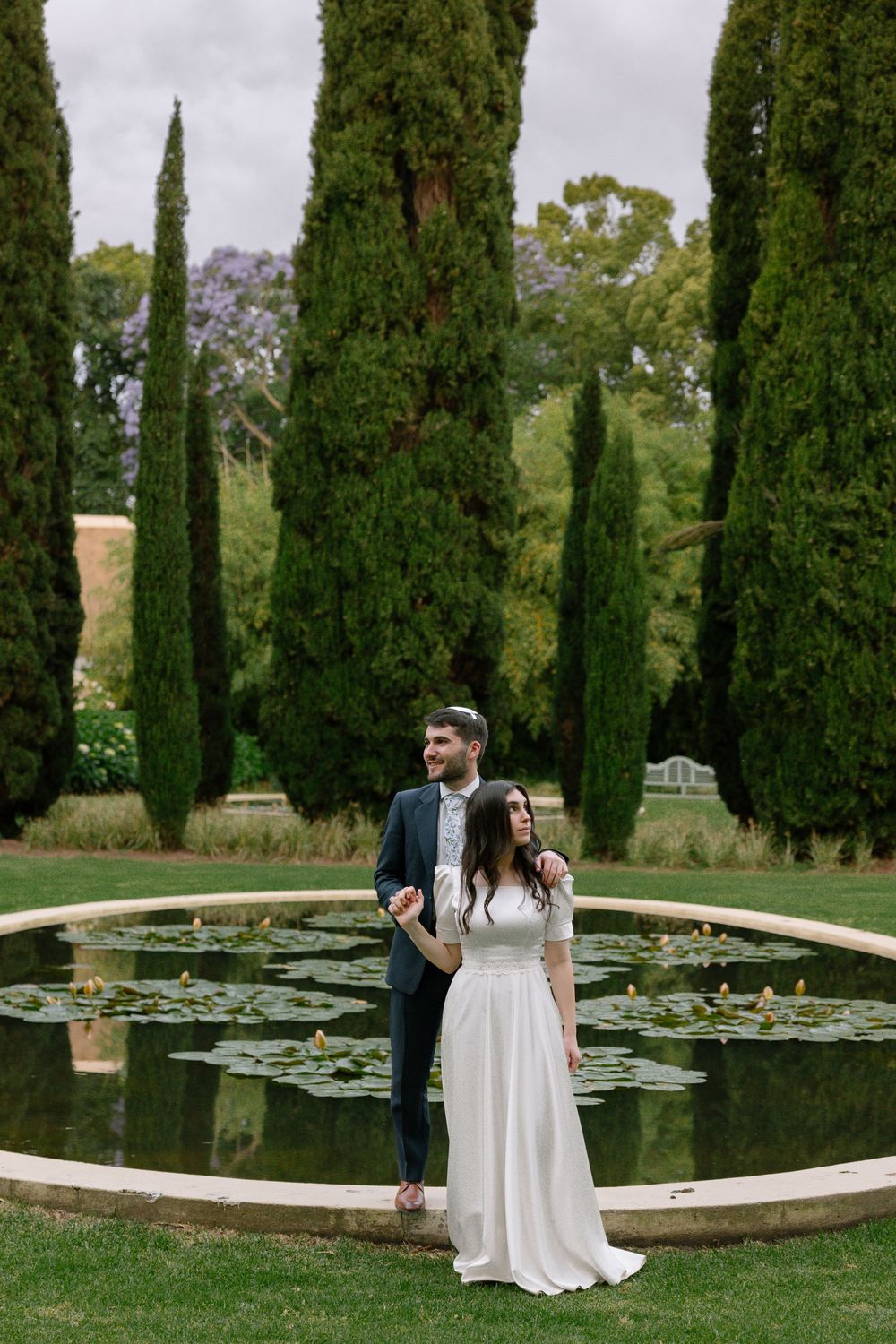 A couple stands by a circular lily pond surrounded by tall cypress trees in an elegant garden setting.