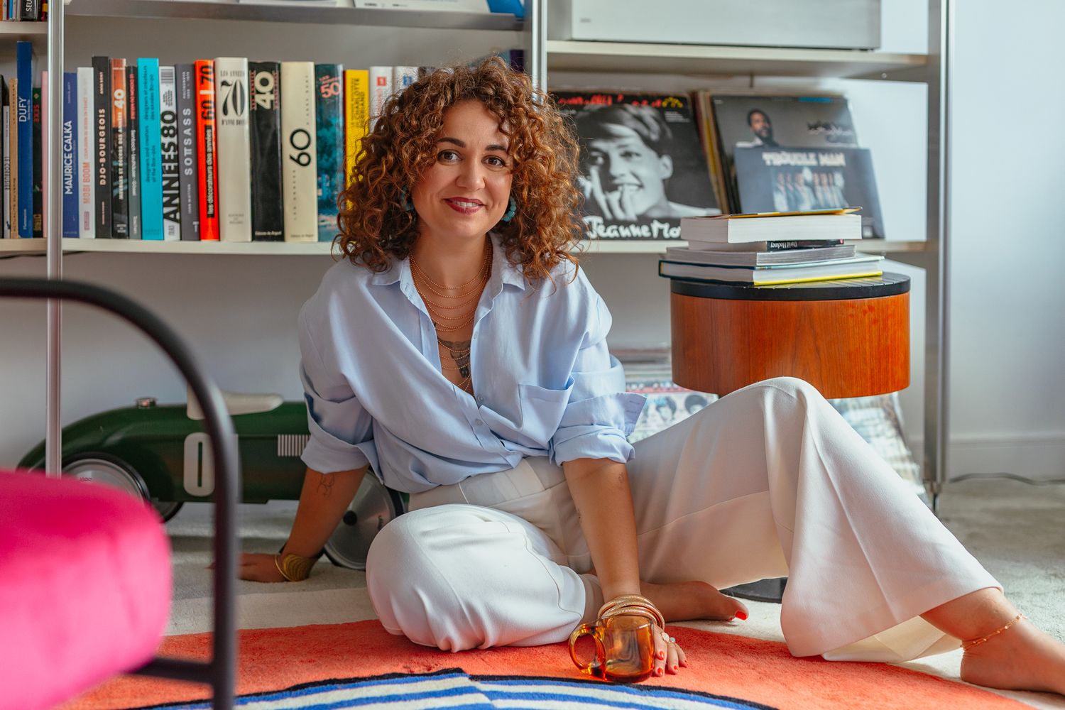 Woman with curly hair sitting on floor beside bookshelf in bright modern living space.