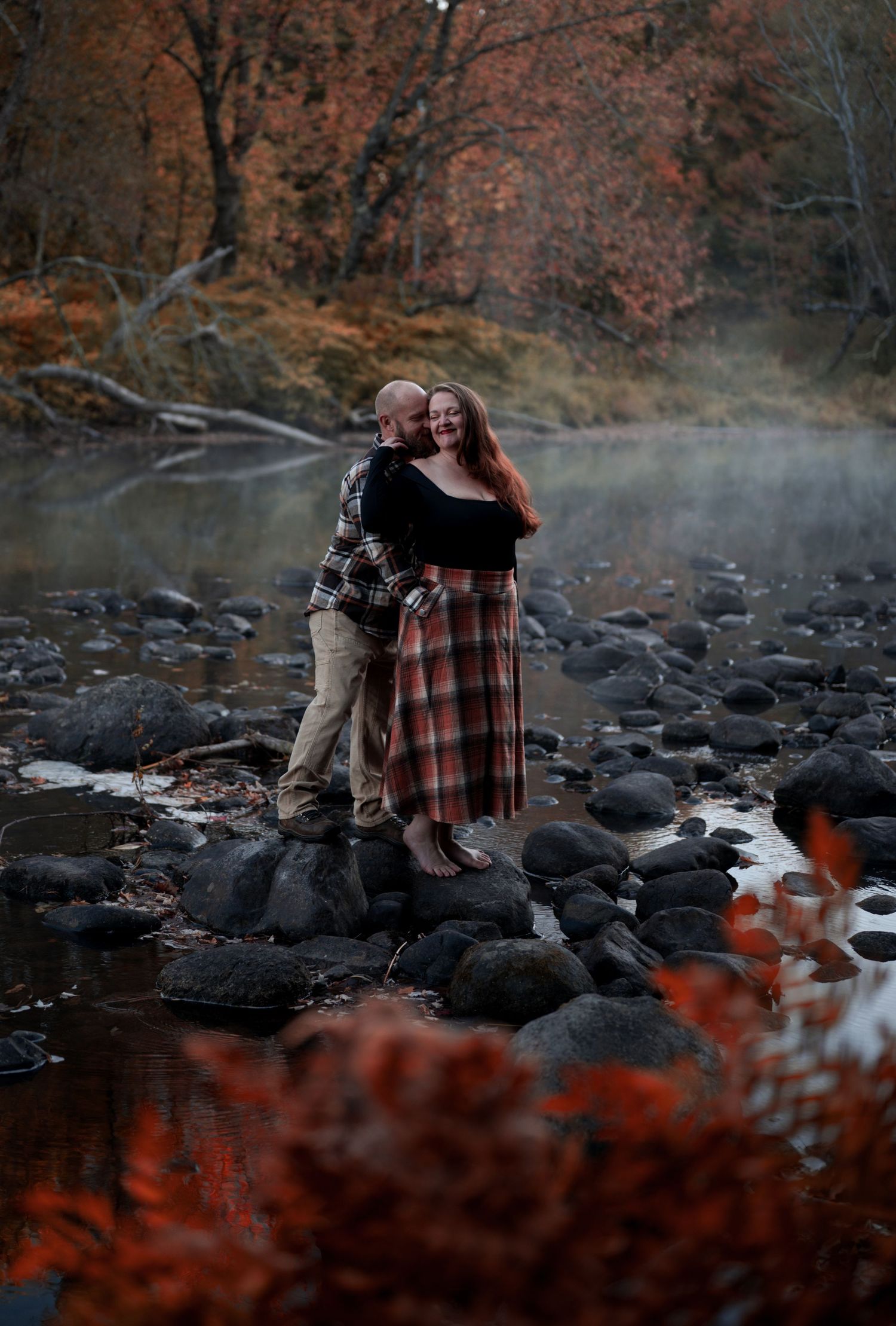 A couple embracing by a misty river surrounded by autumn foliage and dark rocks.