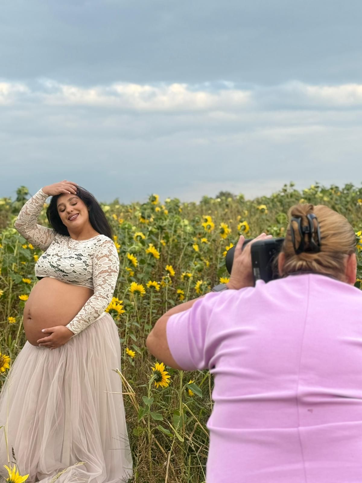 A photographer captures a maternity photo shoot in a field of bright yellow wildflowers on a cloudy day.
