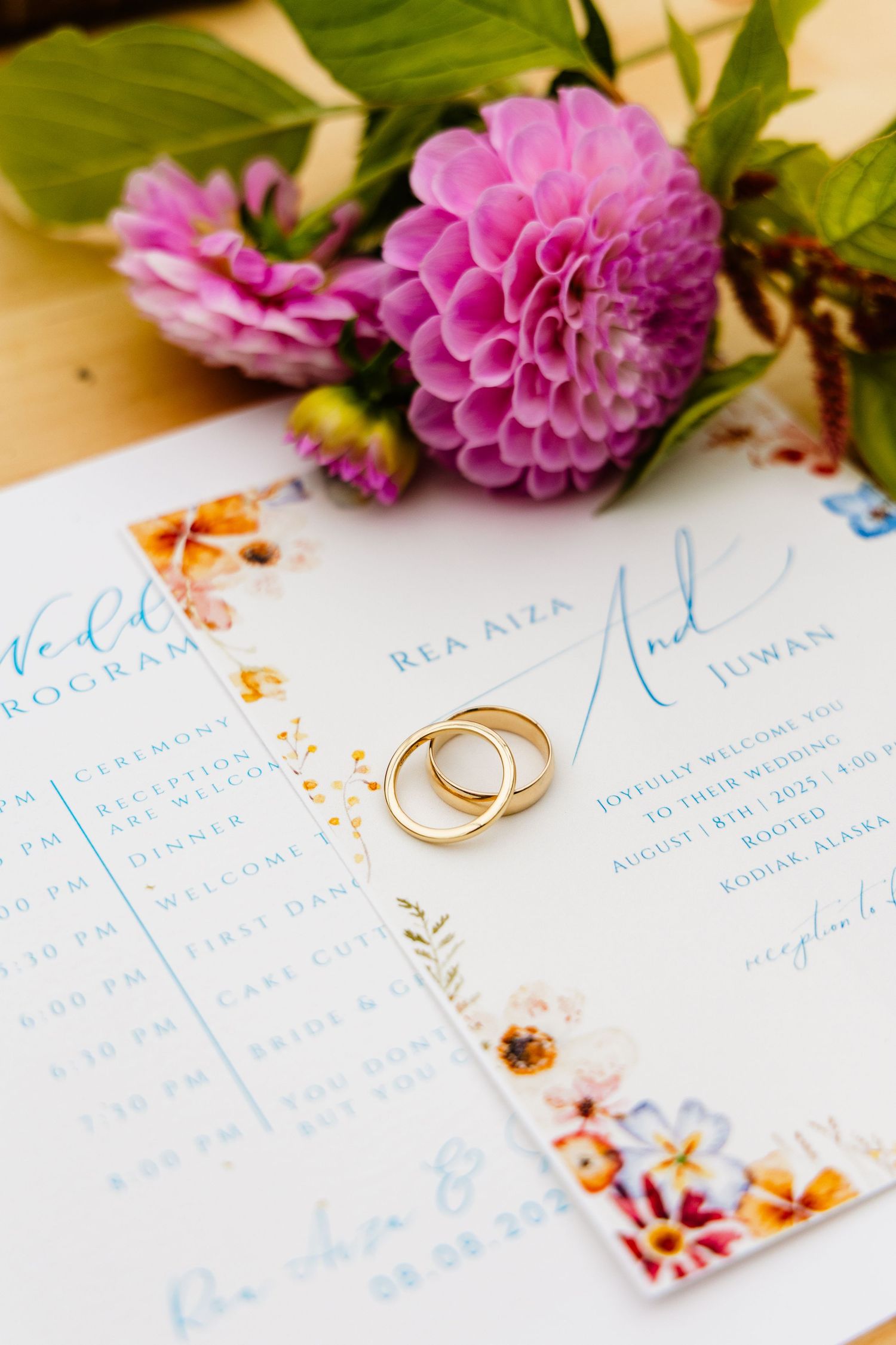 Wedding invitation suite with pink dahlia flowers and gold wedding rings on white paper with blue calligraphy details.
