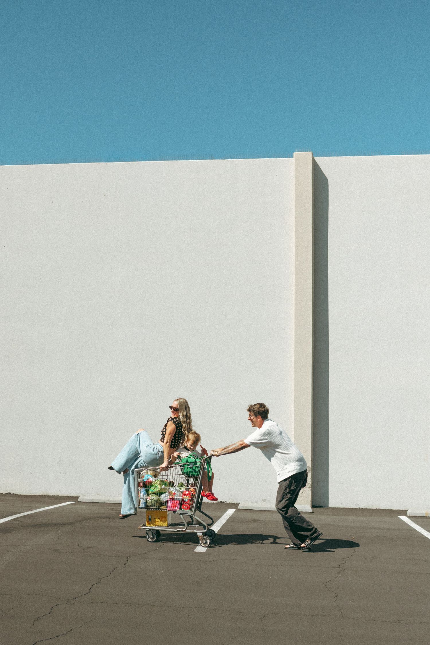 Skateboarders practice tricks against a minimalist white wall backdrop under bright blue sky.