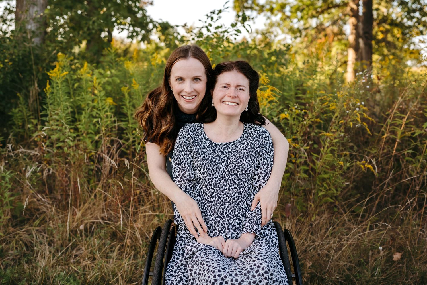 Two people embrace while sitting outdoors in a natural setting with golden sunlight and wildflowers.
