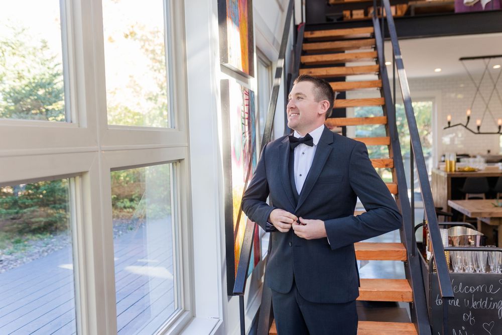 A person in a black tuxedo and bow tie adjusts their jacket while standing near a modern staircase with wooden steps.