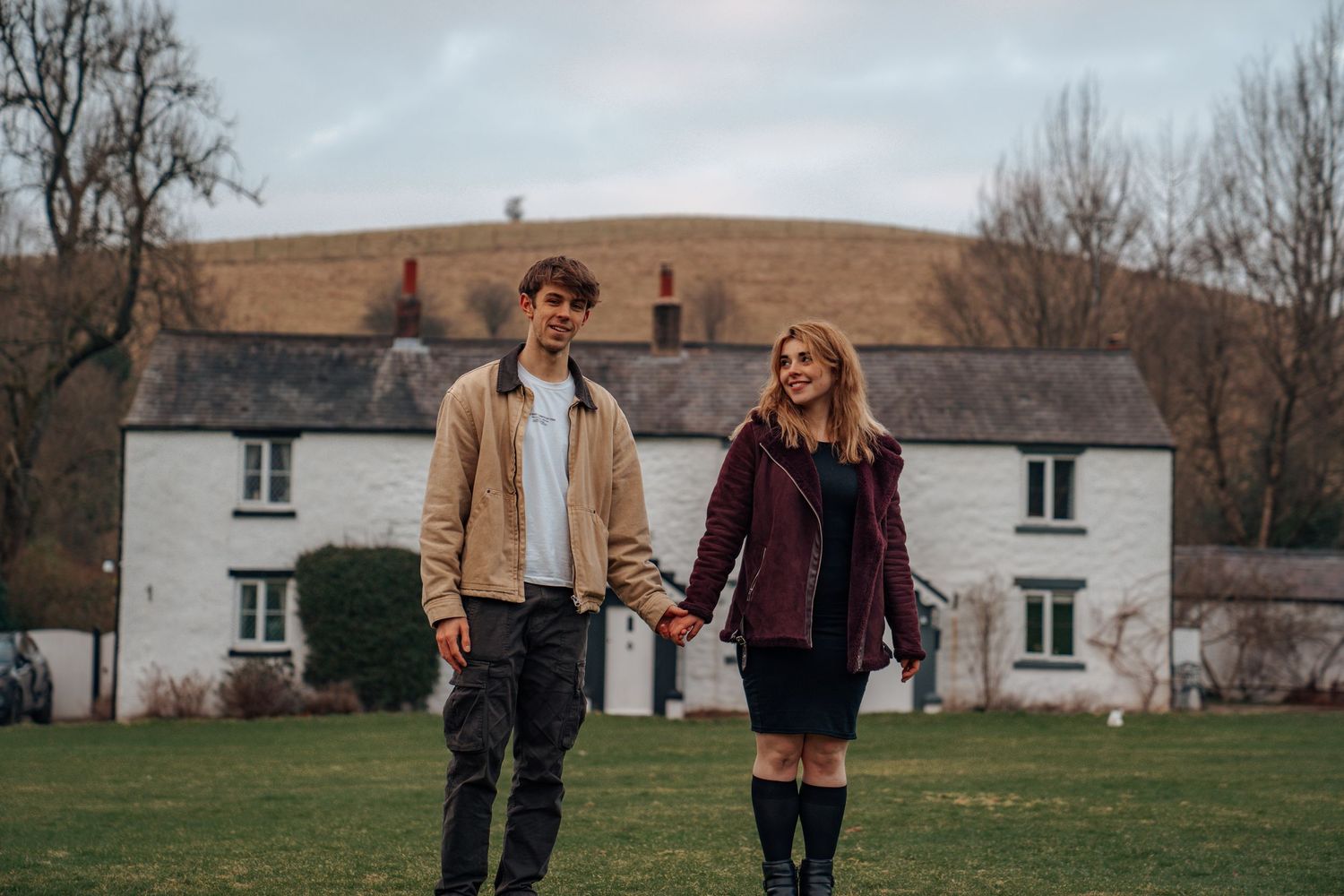 Young couple holding hands while walking across green field with white farmhouse and hills in background.