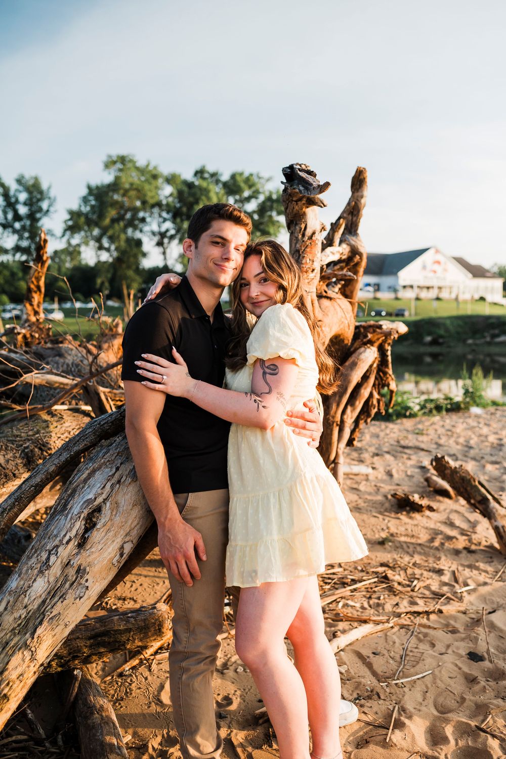 A couple shares a tender moment on a beach with driftwood and trees in the background.