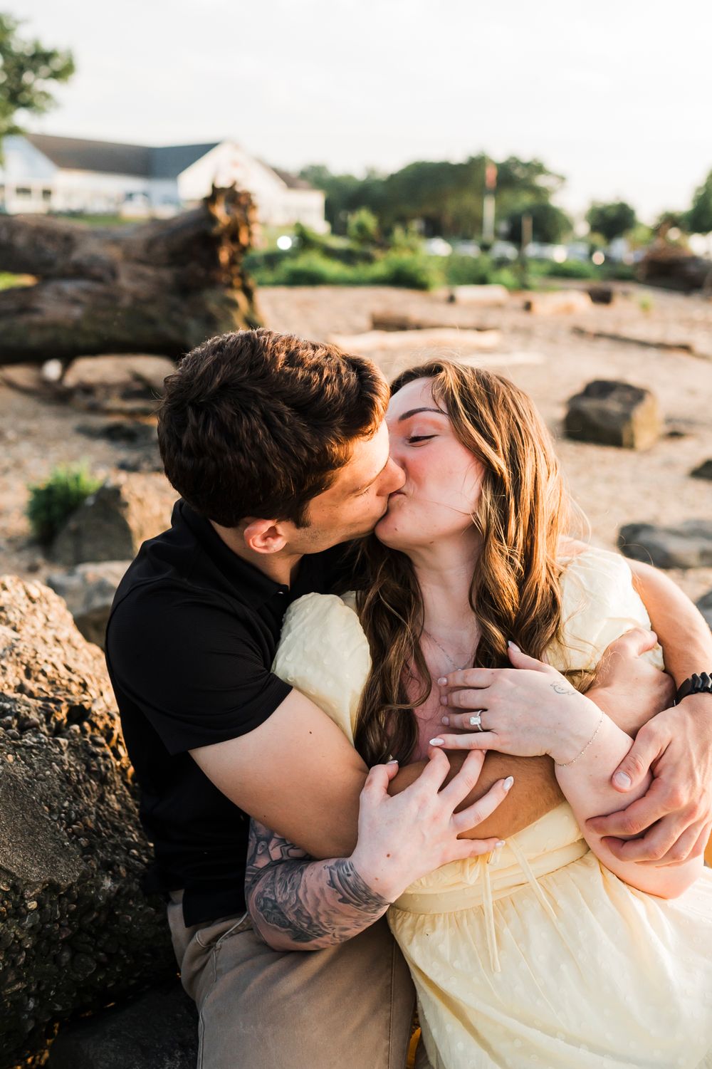 A romantic couple shares intimate kisses while sitting on rocky beach terrain at sunset.