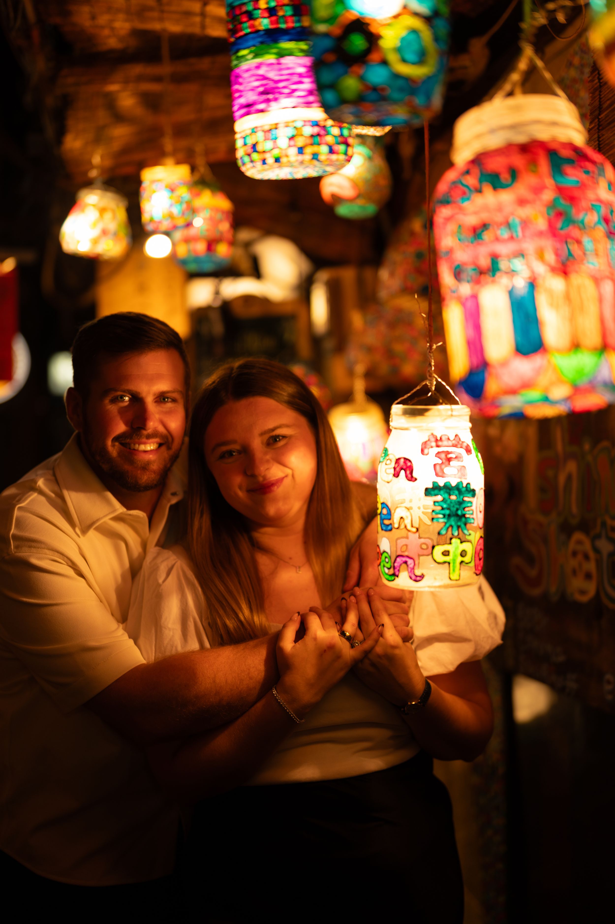 Smiling couple holding a colorful glowing lantern surrounded by hanging lanterns.