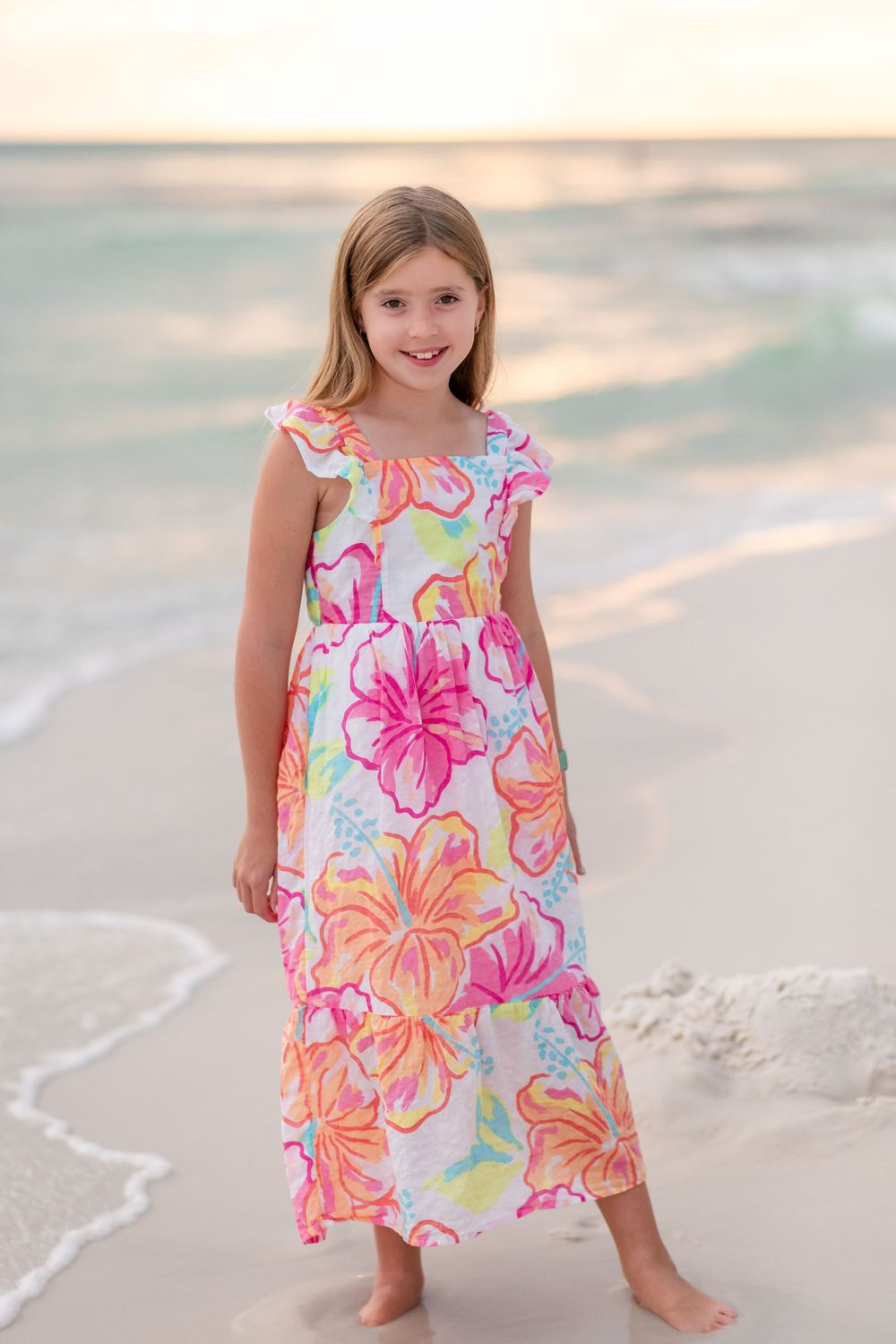 Child in colorful floral dress poses playfully on sandy beach at sunset with ocean waves in background.