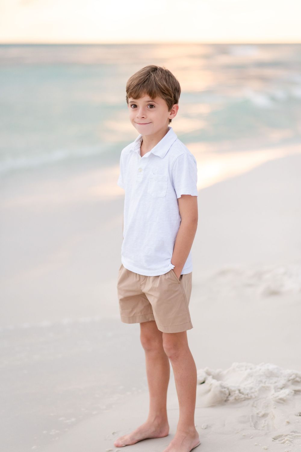 A child in white shirt and khaki shorts stands barefoot on a sandy beach at sunset.