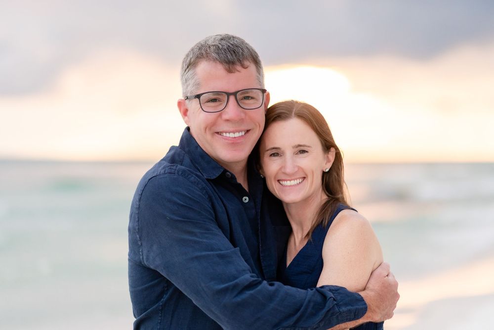 Couple embraces on a beach at sunset with soft pink and orange clouds in the background.