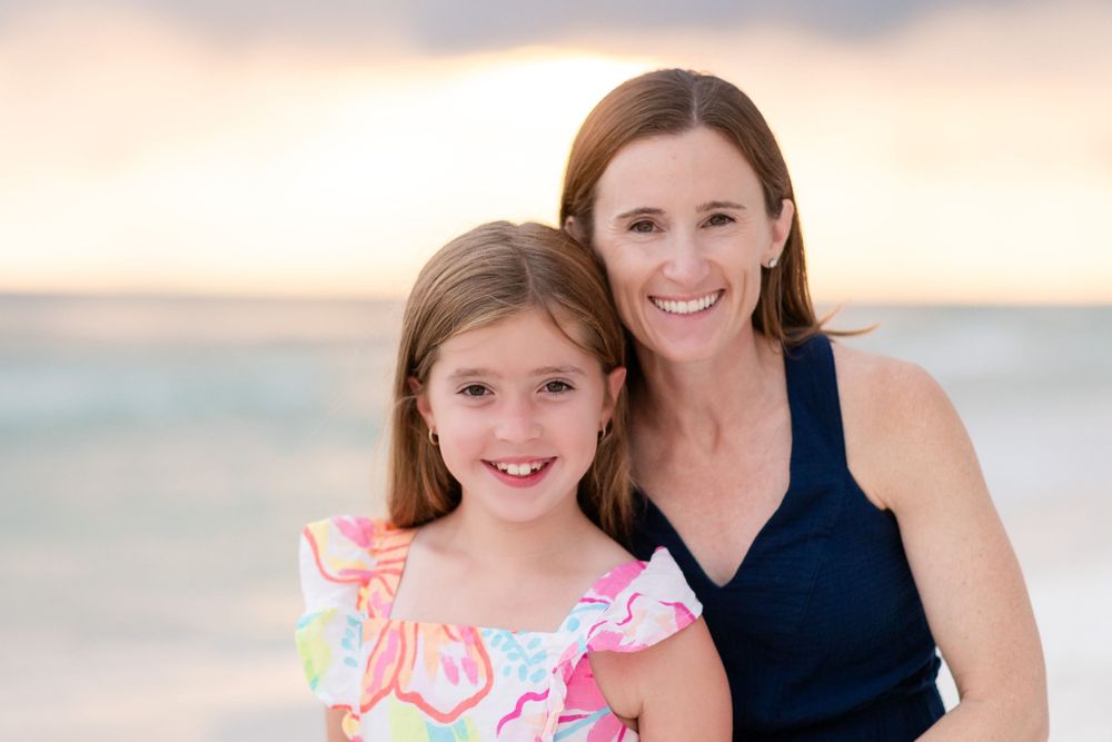 A parent and child pose together on the beach at sunset with warm lighting and soft colors in the background.