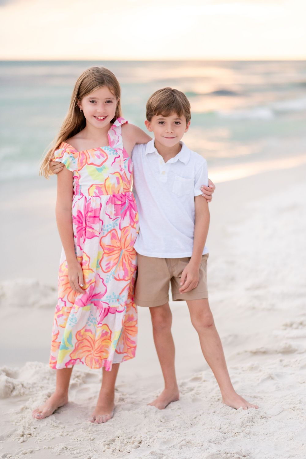 Two children in summer outfits playing together on a white sandy beach at sunset.
