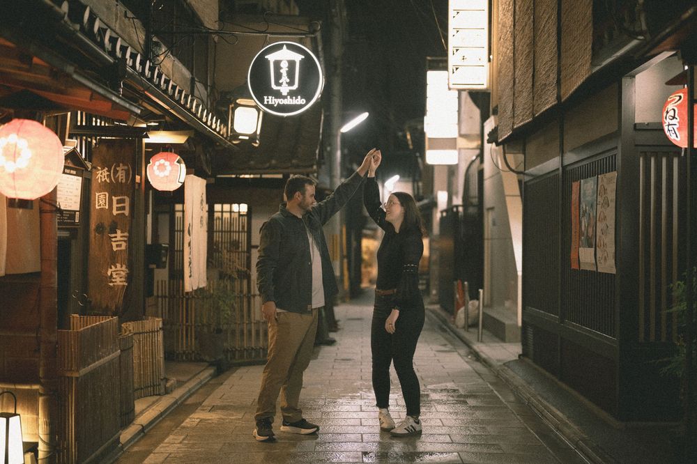 Couple dancing joyfully on a narrow illuminated Japanese street at night with glowing lantern signs.