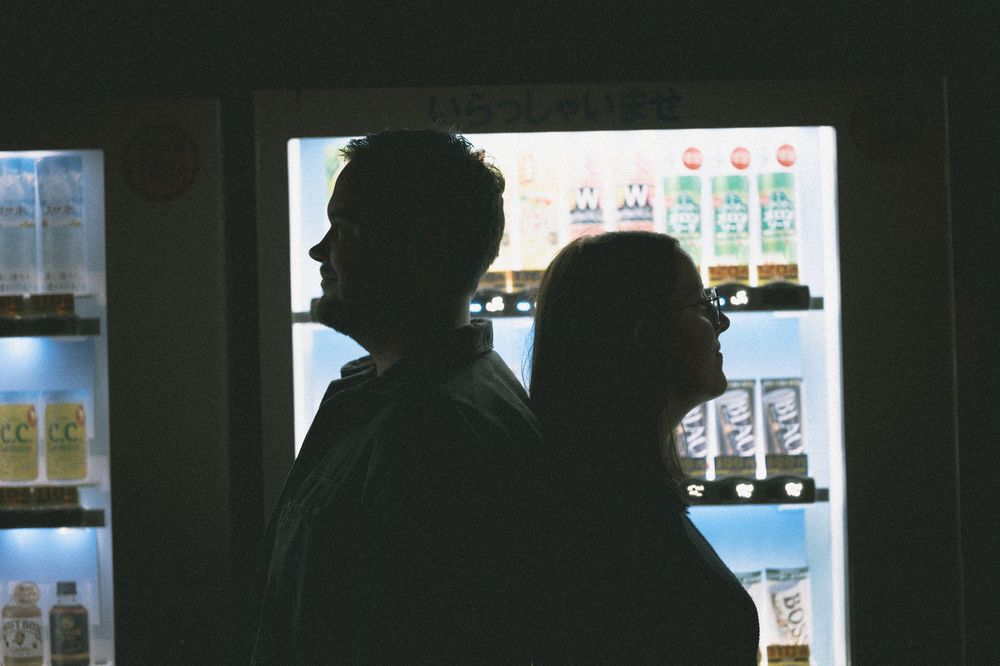 Silhouette of a couple standing in front of a brightly lit Japanese vending machine at night.