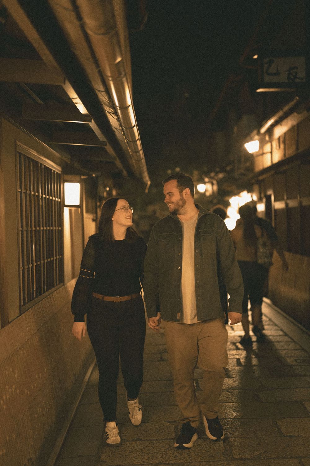 Happy couple holding hands and walking through a narrow lantern-lit Japanese alley at night.