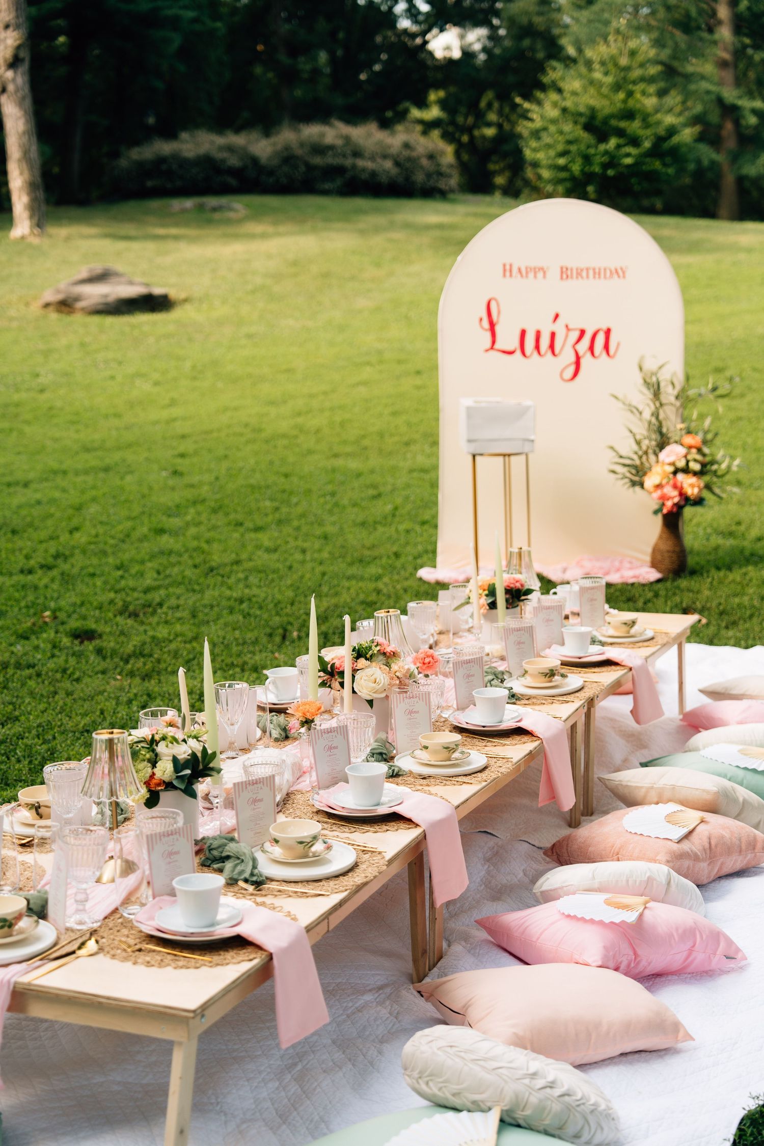 Elegant outdoor picnic setup with low tables, pink cushions, and floral arrangements for a summer garden party celebration.