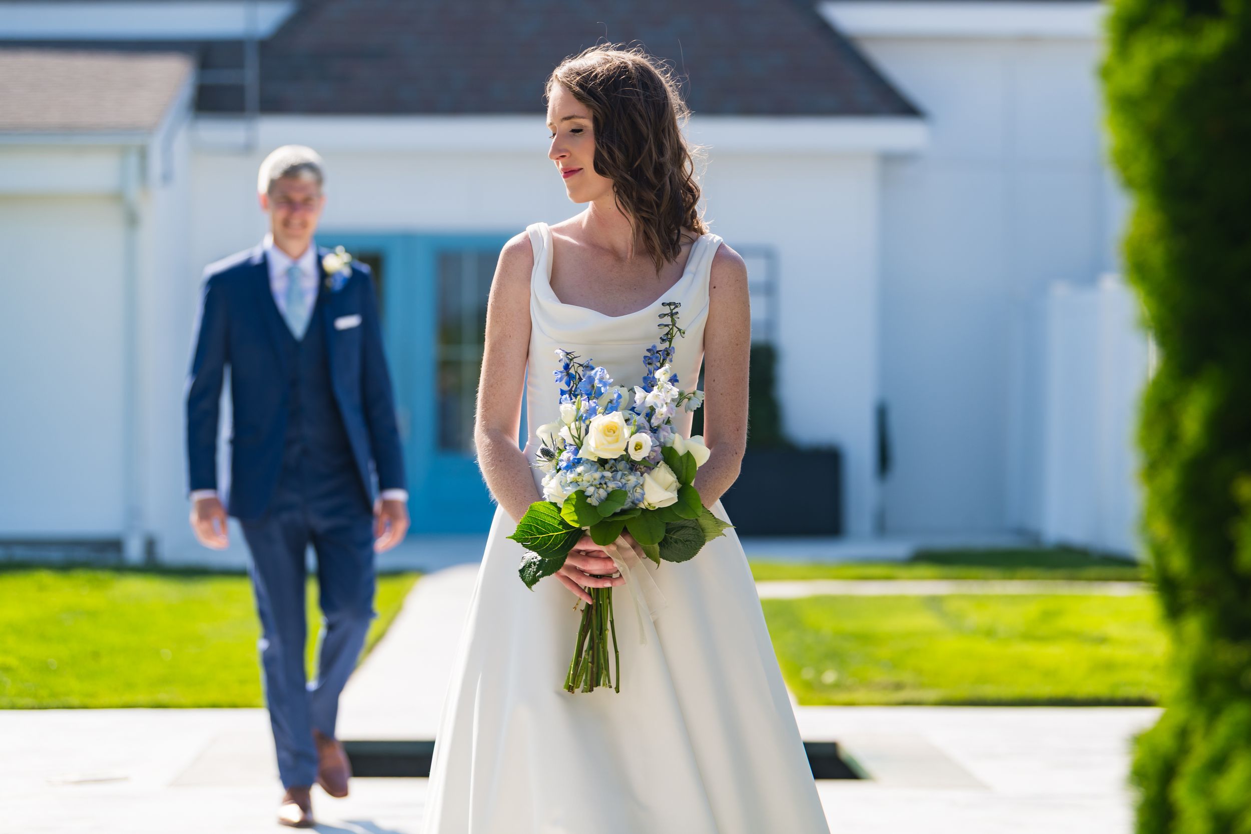 Wedding party members walk and pose together on a sunny day with green lawn and white buildings.