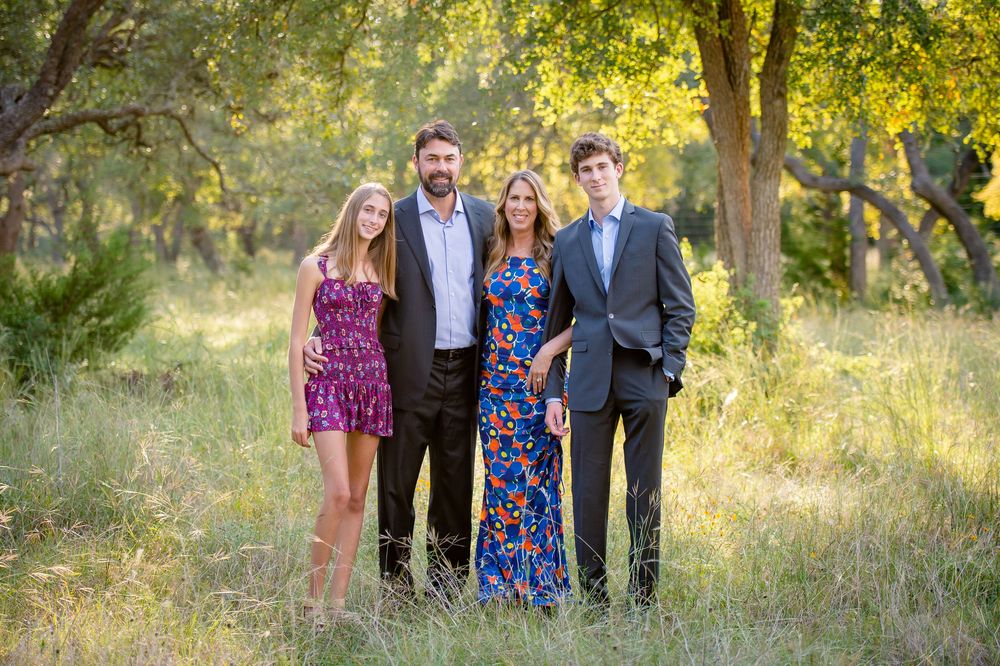 A group of people walking together through a sunlit wooded area wearing casual and formal attire.
