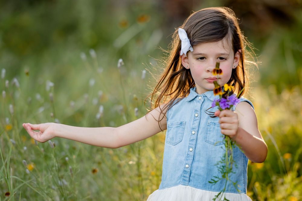 A person wearing a denim vest holds wildflowers while standing in a sunny meadow during golden hour.