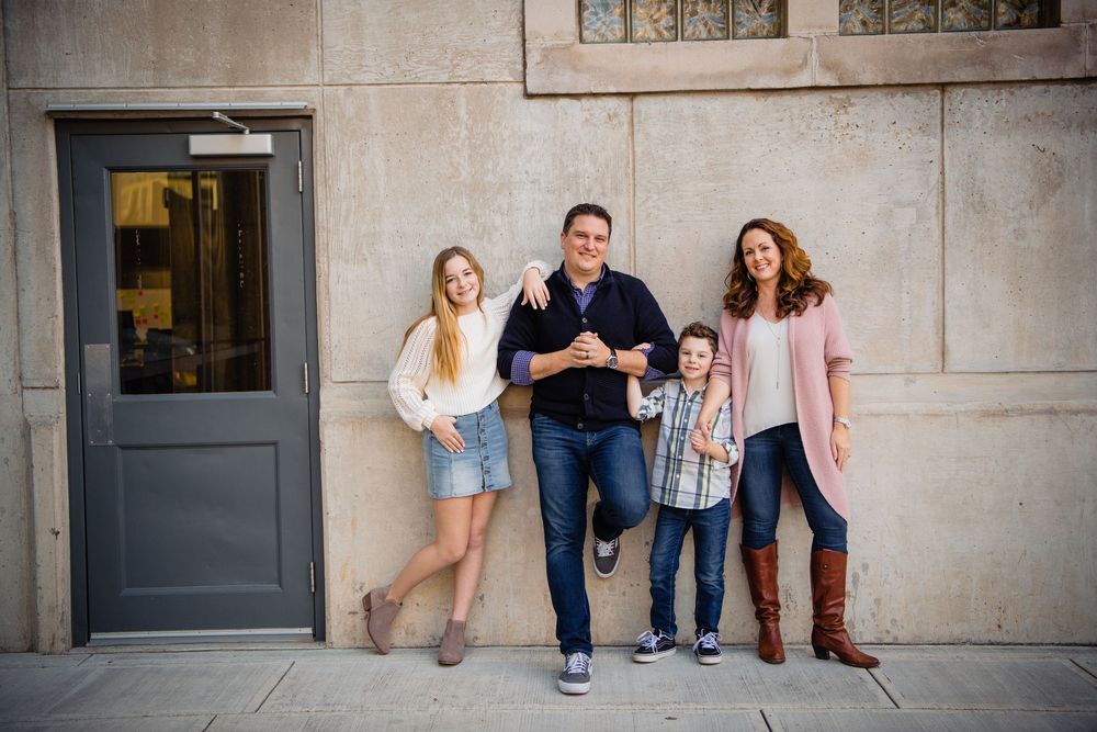 A family in casual clothing sits and poses together against a concrete wall in an urban setting.