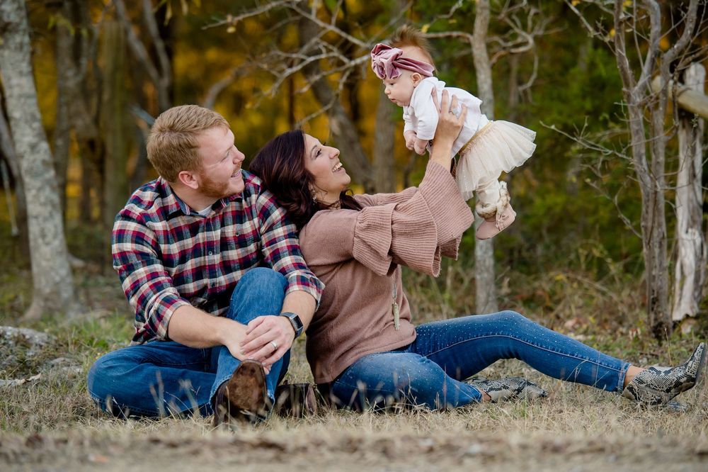 A couple relaxes outdoors with their small white dog in a wooded setting during an autumn photoshoot.