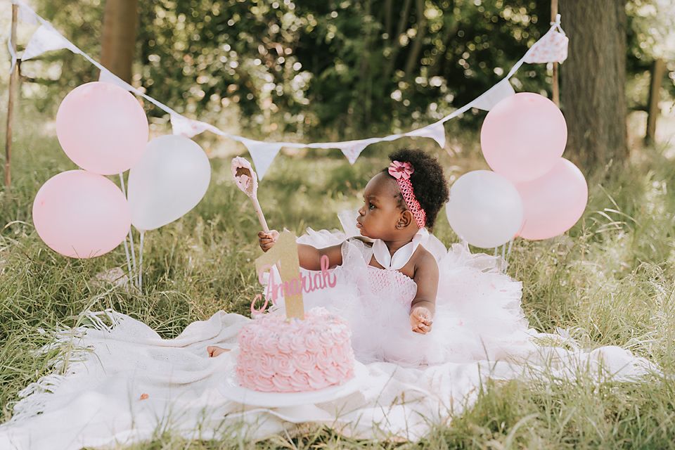 Outdoor first birthday photoshoot with pink balloons and white picnic blanket in garden setting.