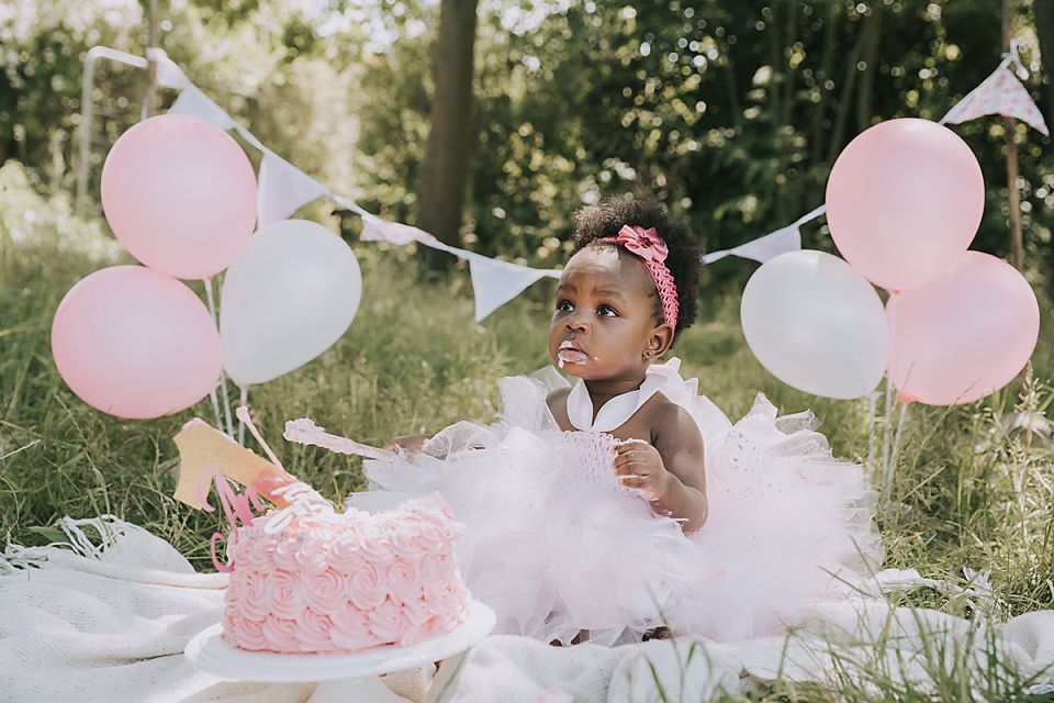 Outdoor first birthday celebration with pink cake and white tulle dress against natural greenery.