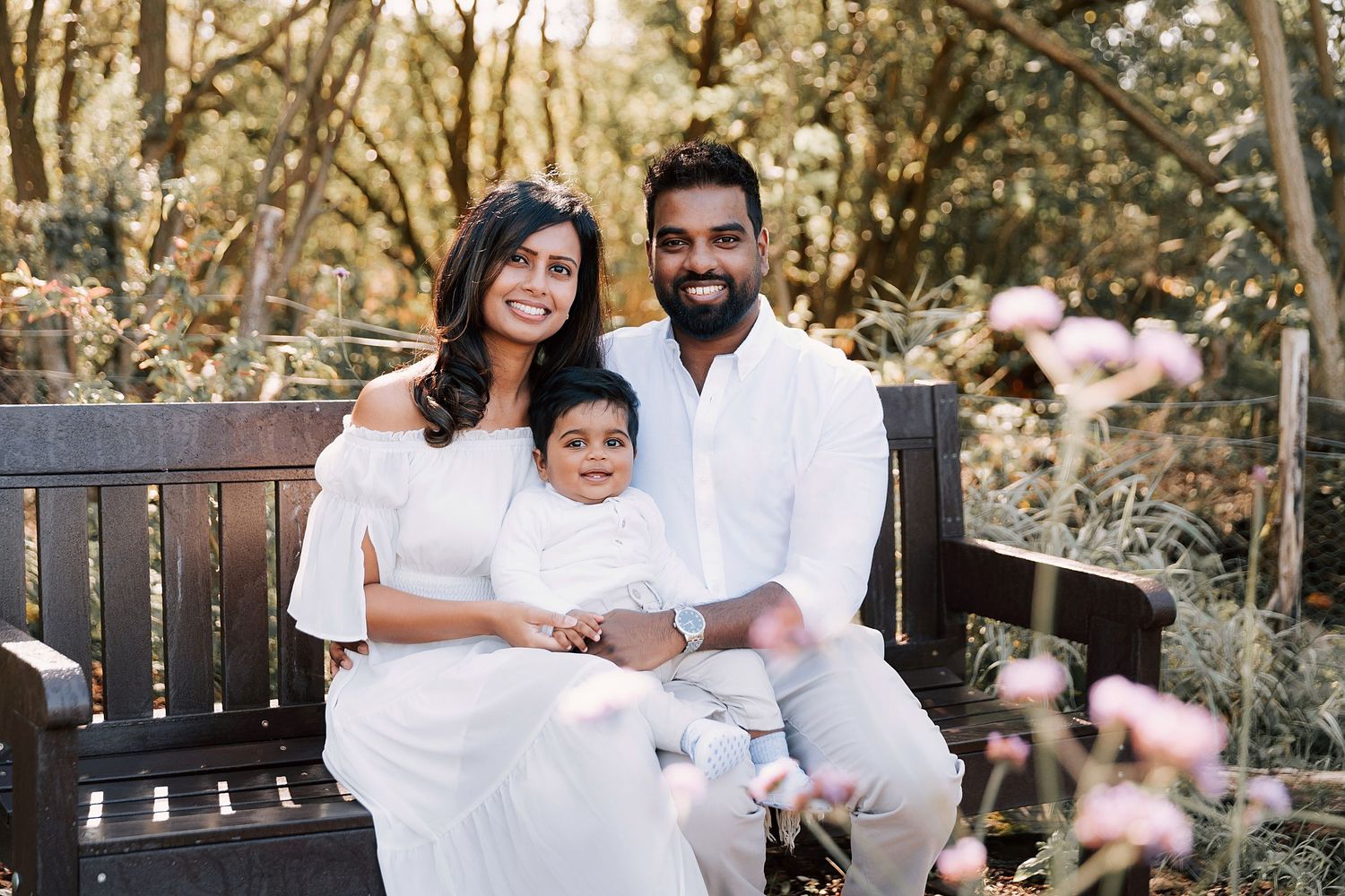 Family relaxing together on outdoor bench surrounded by trees and pink flowers.