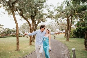 Romantic couple in turquoise and white outfits embrace and walk together on a tree-lined path during a photo session.