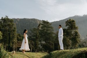 A couple in white attire stands on a grassy hilltop with mountains and trees in the background.