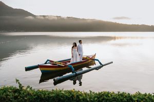 Silhouetted figures paddle wooden canoes across a tranquil lake at sunset with misty mountains in the background.