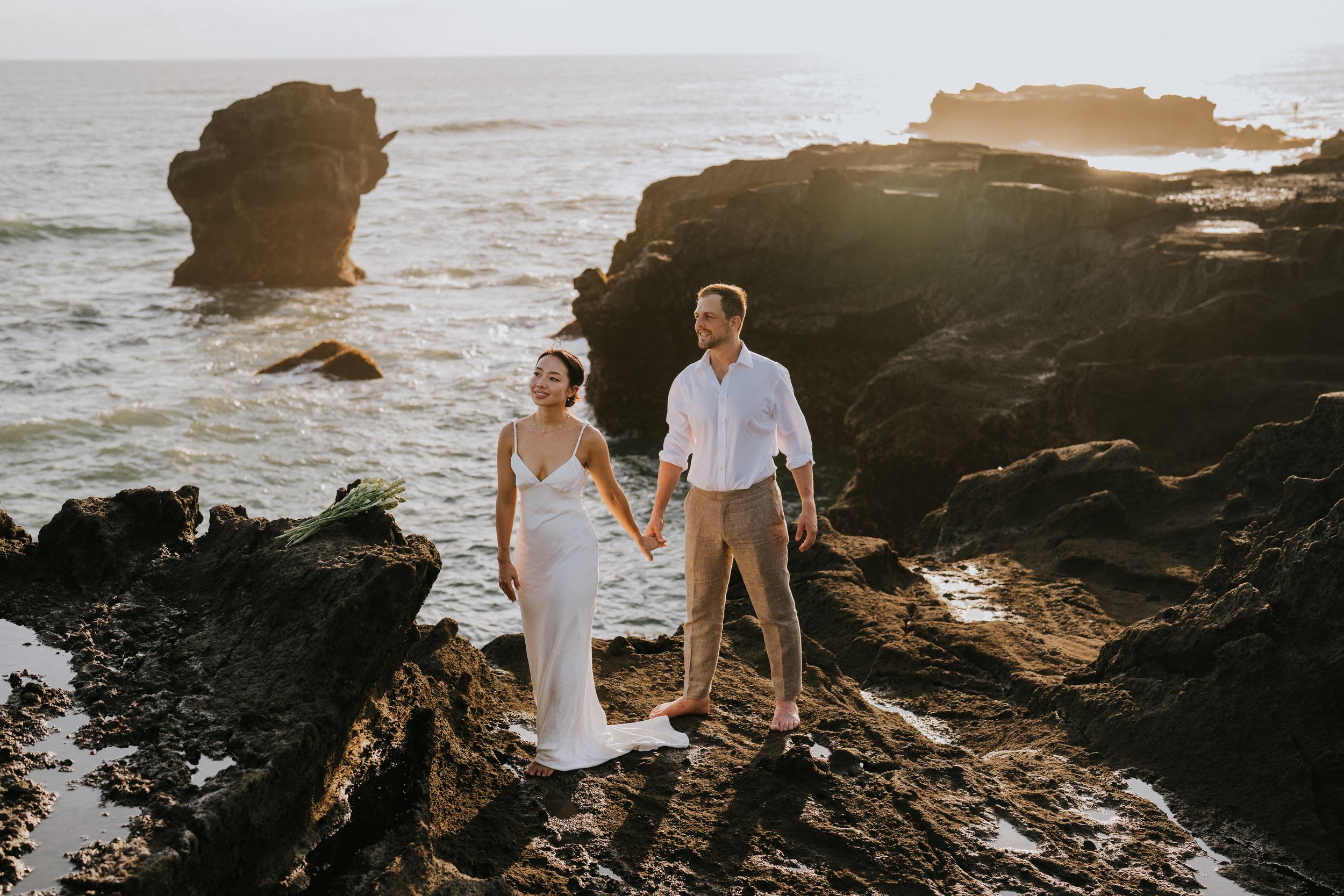 A couple in wedding attire stands on dramatic rocky cliffs at sunset by the ocean.