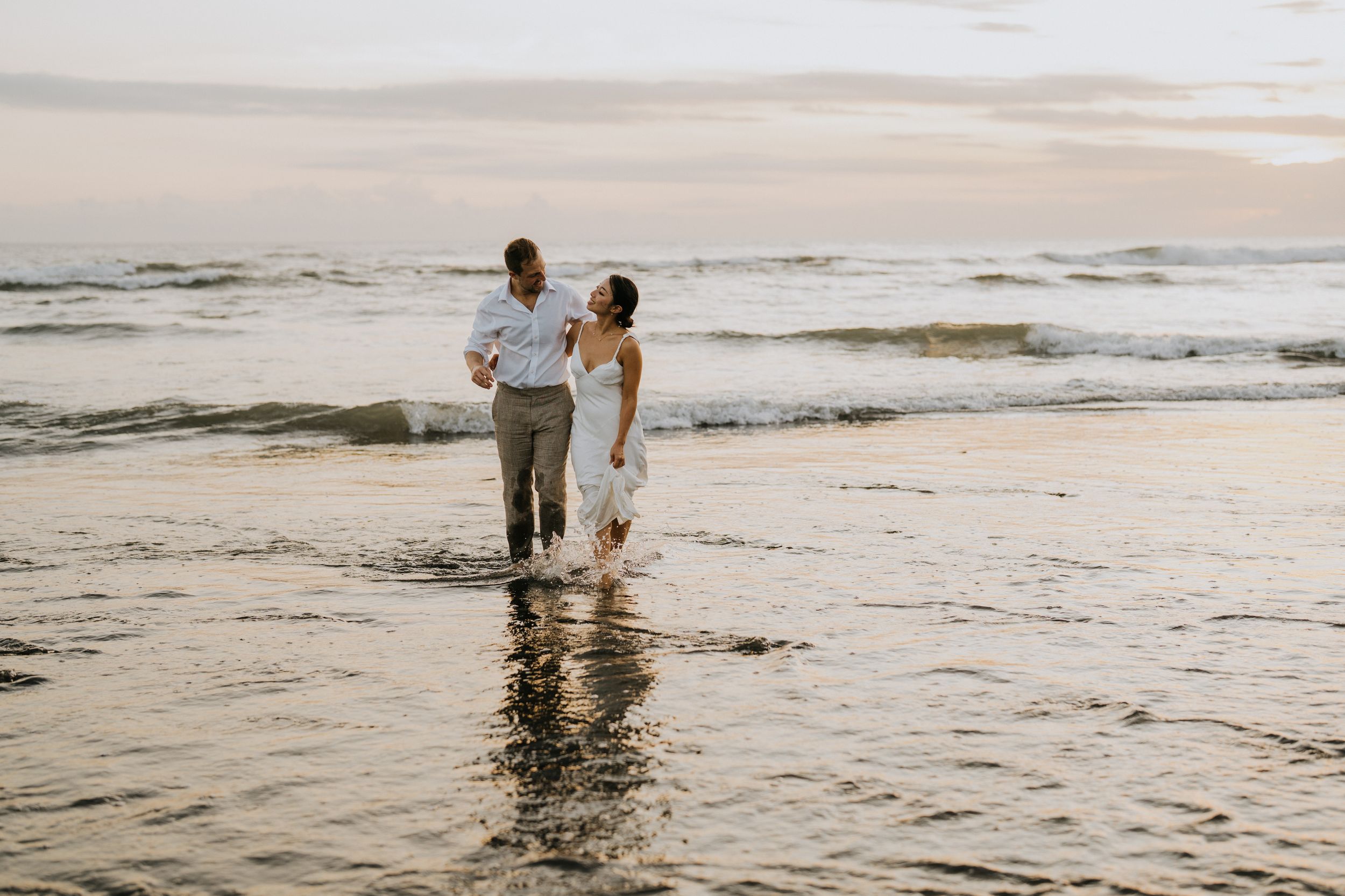 A couple walks together along the shoreline at sunset as gentle waves roll onto the sandy beach.