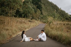 A person in a white dress lies on a winding mountain road surrounded by lush greenery and natural scenery.