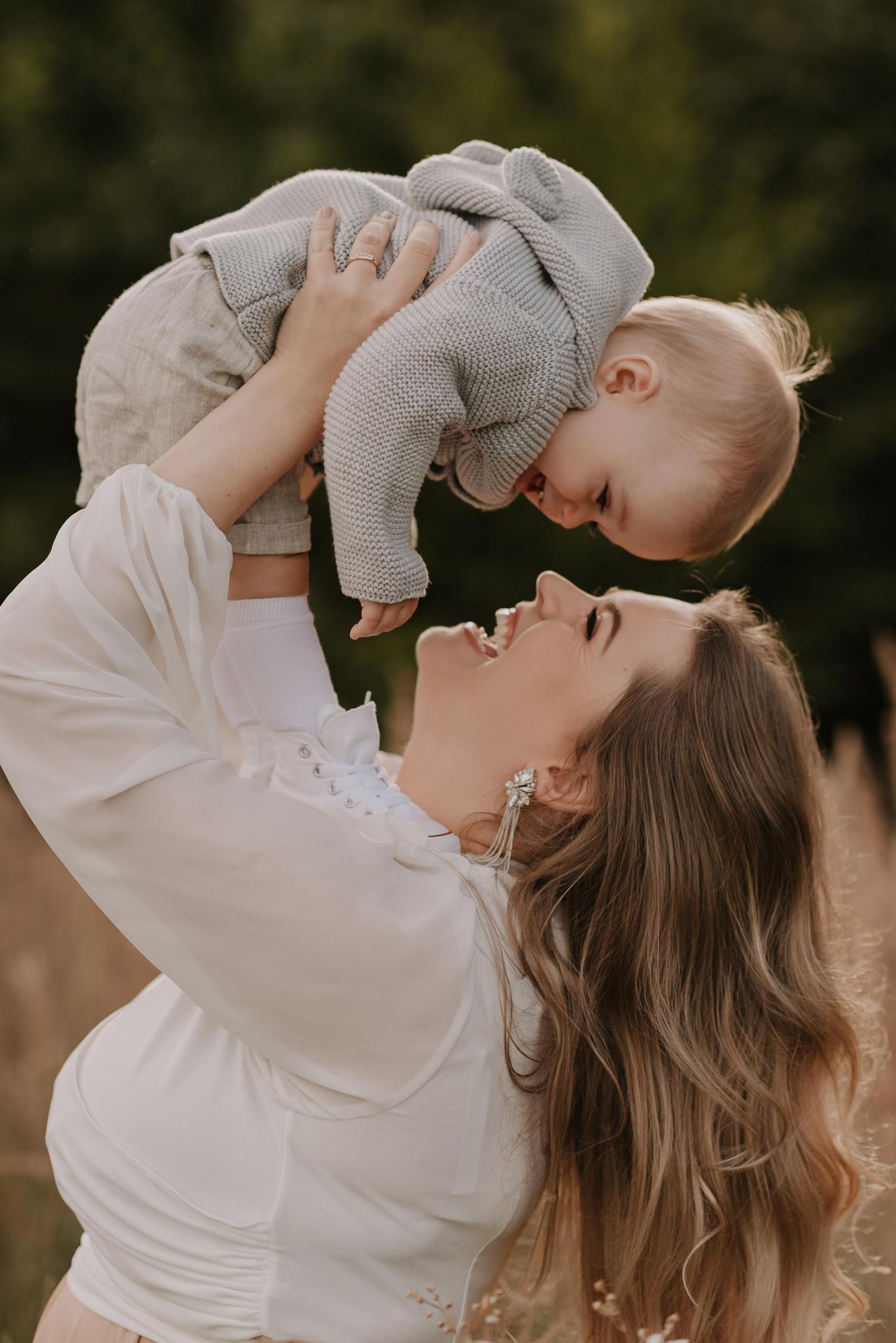 Baby peeks playfully through parent's legs while sitting on grass in a sweet outdoor portrait session.