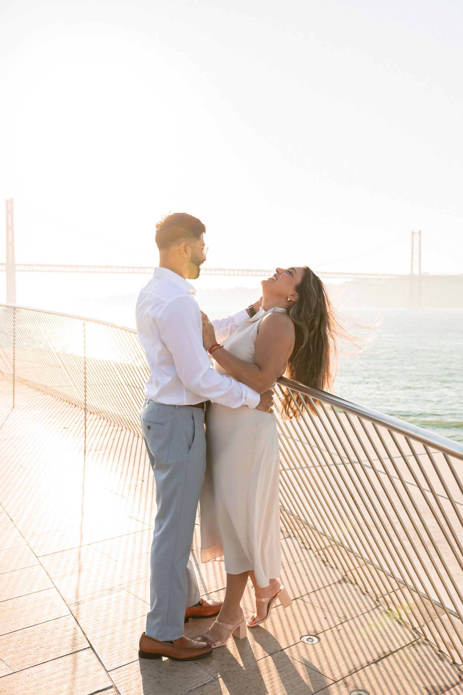 Candid moment of couple laughing together during sunrise engagement session with view of Lisbon’s Bridge. by a Portugal wedding photographer