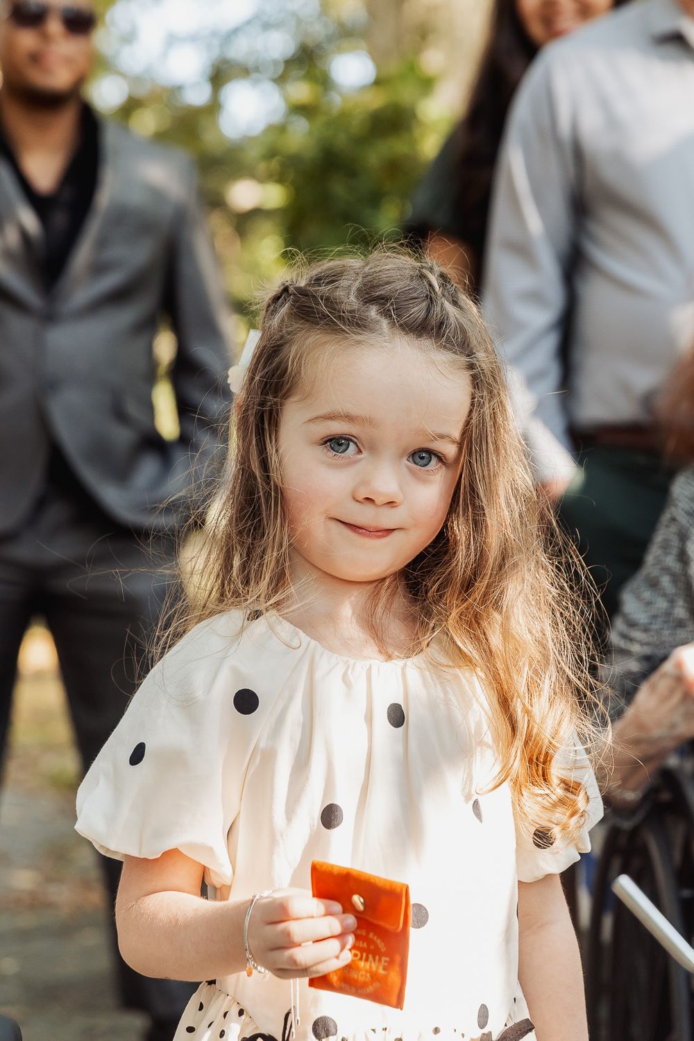 Young child in white dress with puffy sleeves smiles while holding something red outdoors.