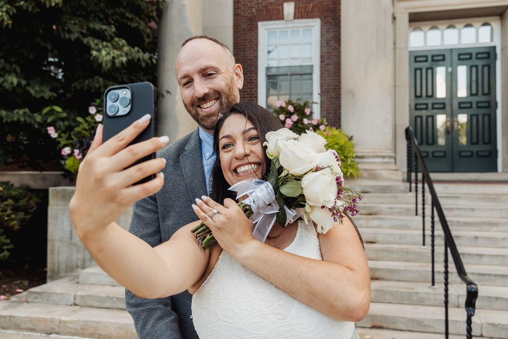 A bride and groom take multiple selfie photos together in front of a stone building.