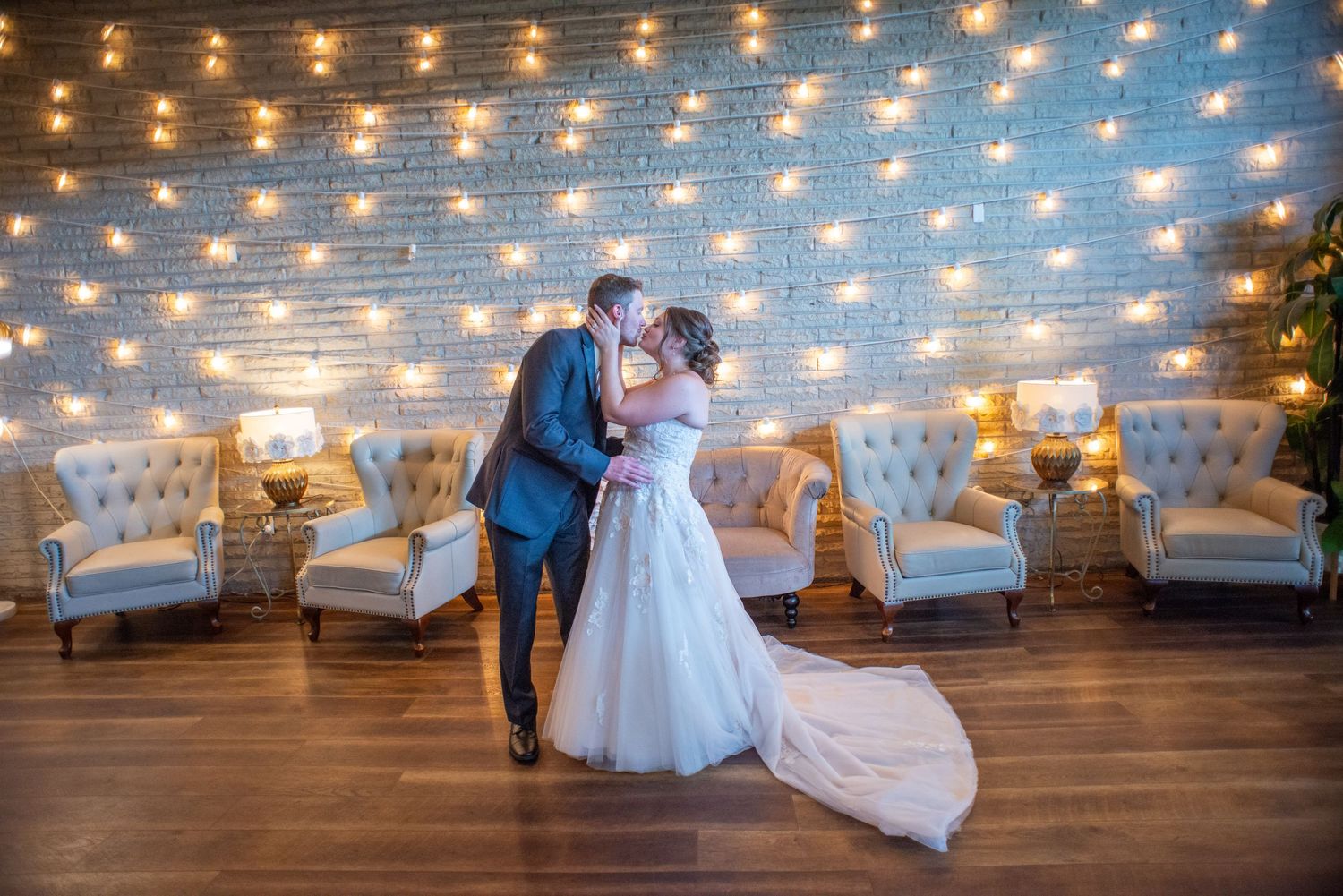 A romantic dance sequence captured under glowing string lights in a rustic indoor venue with white chairs and brick walls.