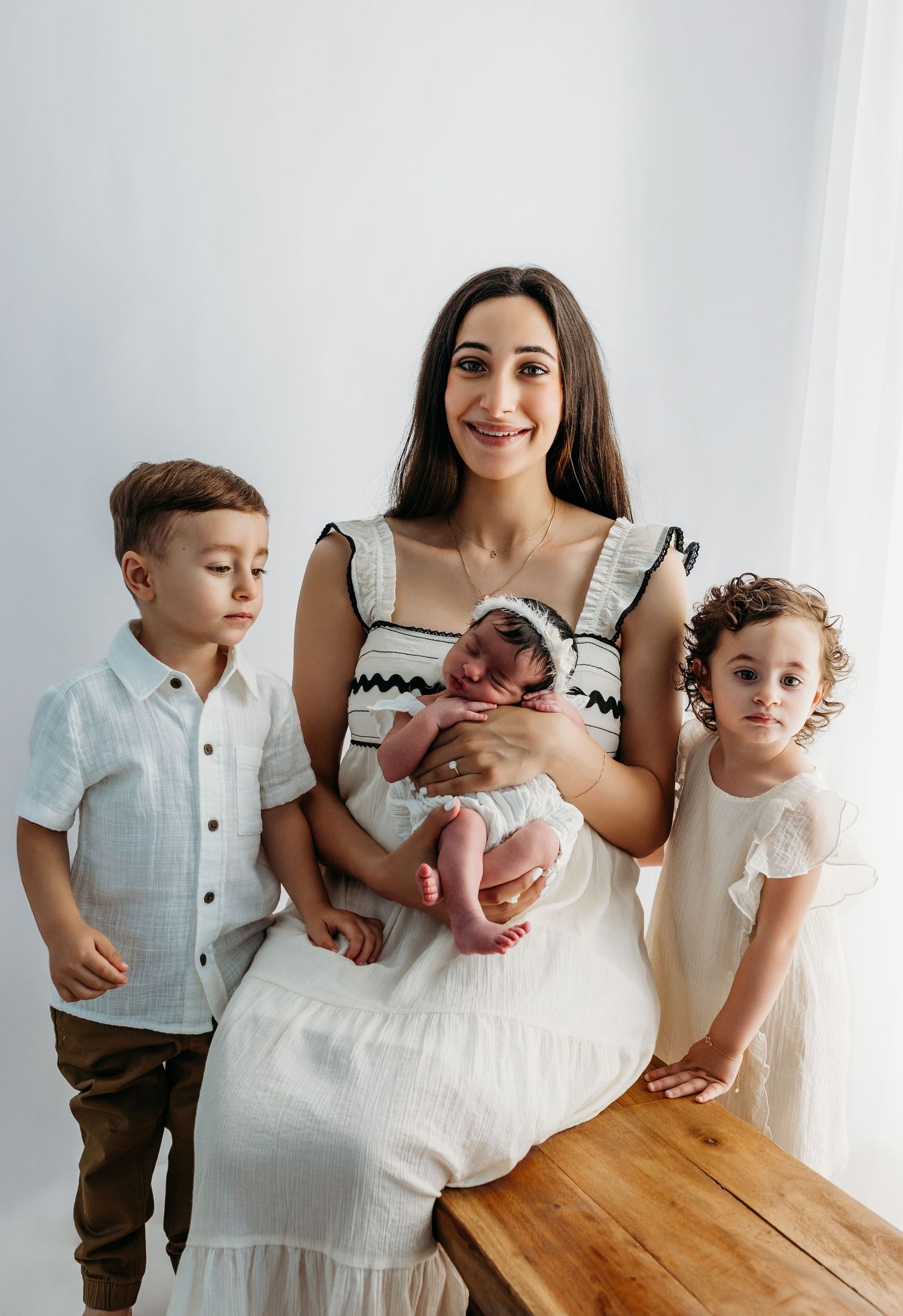Family portrait series showing candid moments with children and a newborn baby around a wooden table in a bright white room.