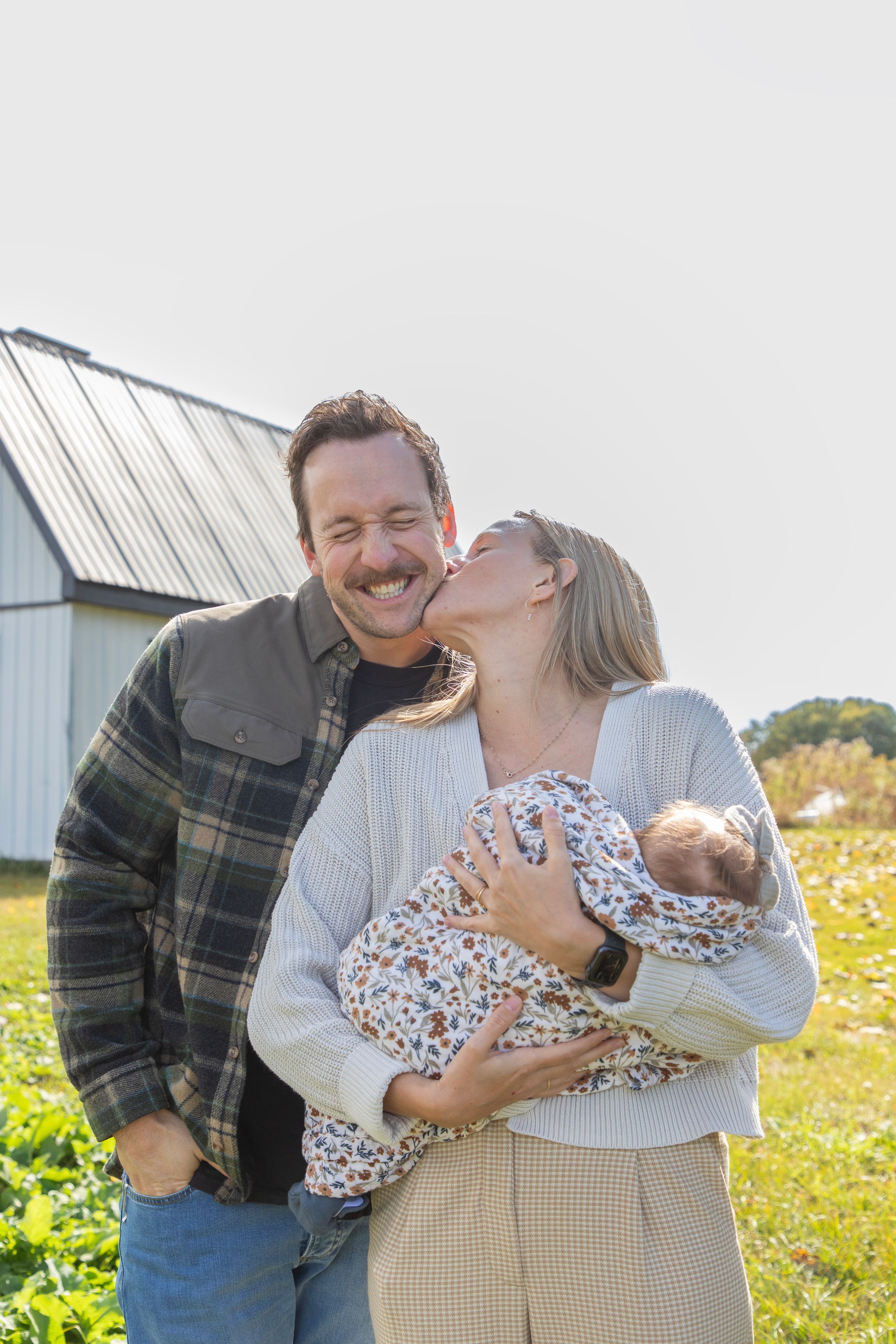 A young family on a farm with the wife in front of the husband, leaning her head back to kiss his cheek. he makes a cheeky smile